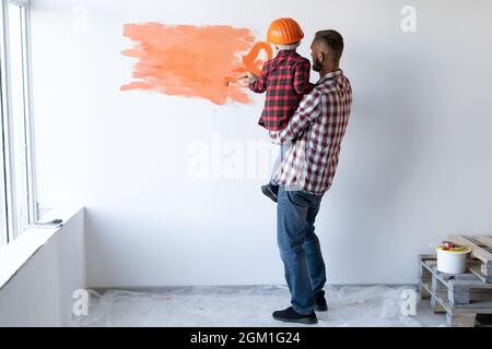 Rückansicht einer Familie, die Hausreparaturen durchführt Vater hält seinen Sohn in den Armen und er malt die Wand orange. Ein Helm auf dem Kopf eines kleinen Malers. Stockfoto