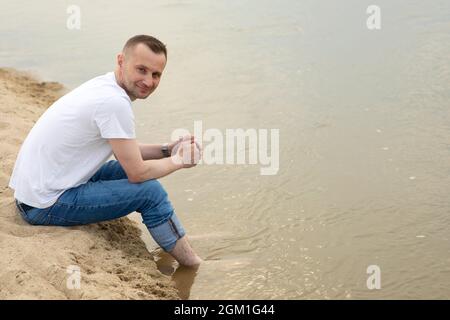 Das Bild einsam positiver und lächelnder Mann, der an einem Strand am Fluss sitzt und seine Beine im Wasser hält Stockfoto