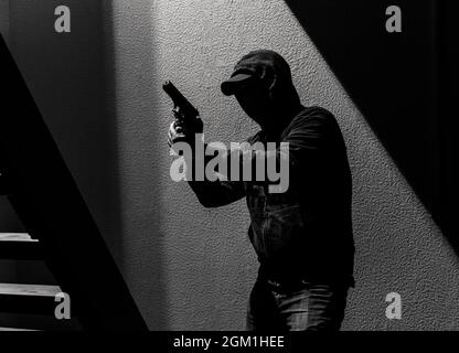 Bewaffneter Verbrecher mit Hoodie und Mütze hält eine Waffe, während er sich durch ein Haus bewegt Stockfoto