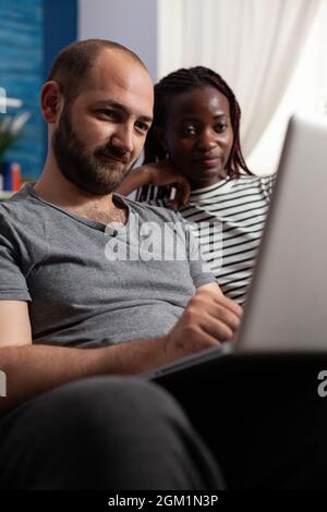 Nahaufnahme eines interracial Paares, das zu Hause auf den Laptop-Bildschirm schaut. Kaukasischer Mann, der an einem digitalen Gerät und einer afroamerikanischen Frau arbeitet, die Display sieht. Multiethnische Menschen mit Technologie Stockfoto