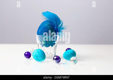 Osterei mit blauer Feder in transparentem Eierhalter auf weißem Tisch. Stockfoto