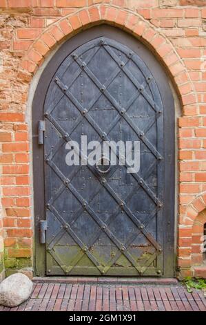 Alte Gebäudefassade mit vintage Eisentür mit kreisförmigem Griff im Zentrum der Altstadt in Litauen. Metall-Grunge-Tor mit Rost und grünem Schimmel in der Backsteinmauer einer Kirche Stockfoto