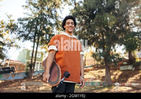 Sorgenfreier Skateboarder mit seinem Skateboard in einem urbanen Park. Fröhlicher Teenager, der die Kamera anlächelt, während er draußen steht. Sportliche Teenager-Tochter Stockfoto