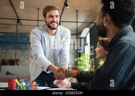 Zwei Männer schüttelten sich die Hände und schauten sich lächelnd an, während ihre Kollegen bei der Geschäftsbesprechung sitzen Stockfoto