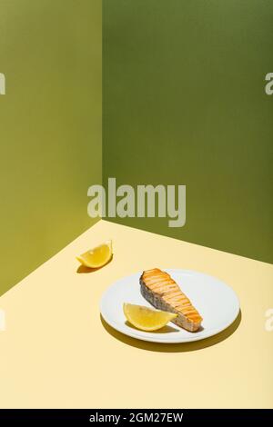 Gebackenes Lachsfilet mit Zitrone auf weißem Teller, hartem, modernem schattengrünem Hintergrund Stockfoto