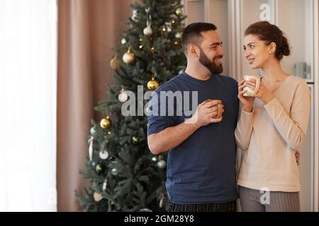 Lächelndes, liebevolles junges kaukasisches Paar, das am Weihnachtsbaum steht und sich umarmt, während es am Neujahrmorgen gemeinsam Kaffee trinkt Stockfoto