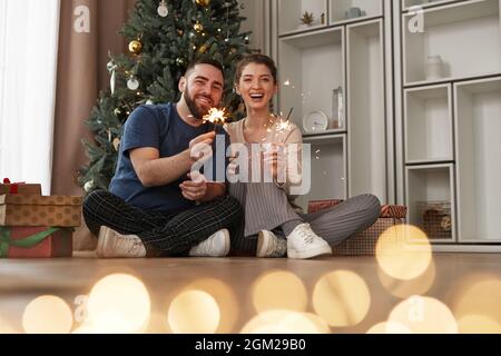 Fröhliche schöne junge kaukasische Paar in gemütlicher Kleidung sitzen gegen Weihnachtsbaum und halten brennende Wunderkerzen zu Hause Stockfoto