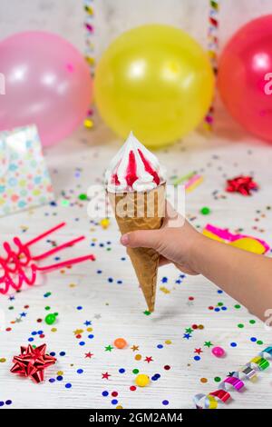 Eiskegel in der Hand eines Kindes auf einem Geburtstaghintergrund mit farbenfrohem Dekor und Süßigkeiten mit verschwommenem Hintergrund. Selektiver Fokus. Stockfoto