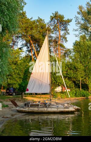 07.17.2021 Oskolsee, Ukraine. Ein Katamaran mit weißen Segeln steht am Ufer in den Strahlen der untergehenden Sonne. Stockfoto