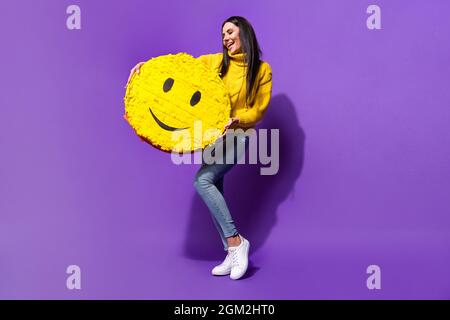 In voller Länge Porträt von zufriedenen jungen Mädchen aussehen Arme halten große Emoticon isoliert auf lila Farbe Hintergrund Stockfoto