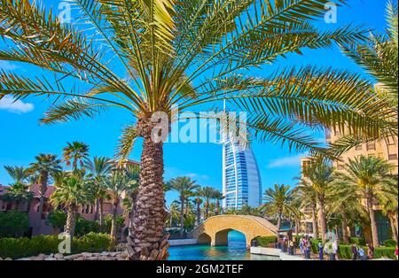 Der Blick auf den Kanal, die kleine Brücke, die Windfänger des Souk Madinat Jumeirah Marktes und das Burj Al Arab Hotel durch die Palmenzweige, die sich im Wind wiegen, D Stockfoto