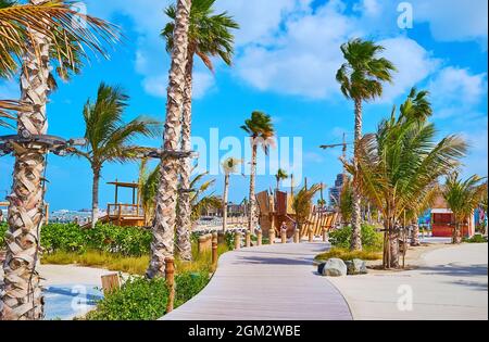 Genießen Sie einen Spaziergang entlang der verwinkelten hölzernen Fußgängerbrücke des öffentlichen Strandes von La Mer, gesäumt von Palmen und üppigen Pflanzen, Dubai, VAE Stockfoto