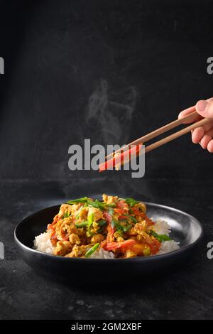 Thailändisches Gericht Gai Pad Pongali mit Huhn, Eiern, würziger, gelber thai-Paprikapaste, Tomaten, Reis in schwarzer Schüssel im Wok. Nahaufnahme. Traditionelle lokale Pause Stockfoto