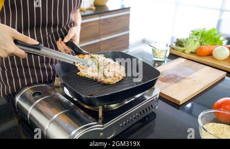 Der Koch verwendet Zangen, um das Steak aus der heißen Pfanne zu zangen. Auf der Theke steht das Holzbrett mit verschiedenen Gemüsesorten. Stockfoto