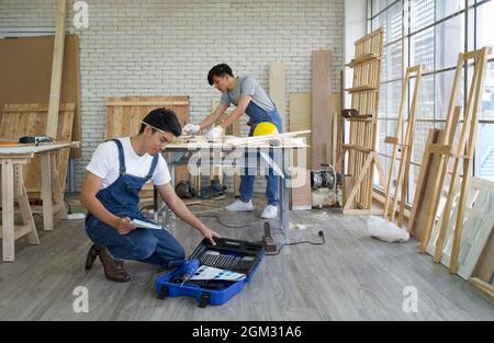 Der junge asiatische Zimmermann saß mit der Ausrüstung selbstbewusst in seiner Werkstatt. Ein anderer Mann, der mit einem Bleistift Zeichnungen auf ein Stück Holz zeichnet. Stockfoto