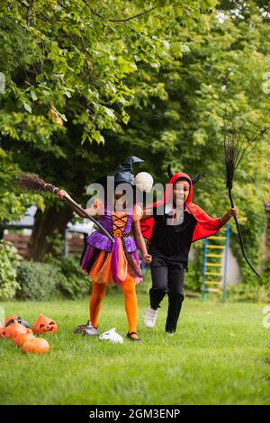 Fröhliche afroamerikanische Kinder in halloween-Kostümen halten Besen auf dem Hinterhof Stockfoto