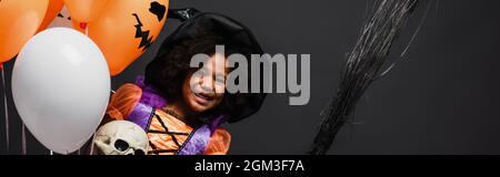 Happy african american Girl in halloween Kostüm hält Besen und Ballons isoliert auf schwarzem, Banner Stockfoto