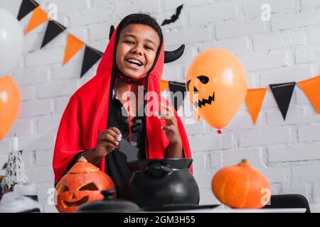 Glücklicher afroamerikanischer Junge im Teufel halloween Kostüm, der papiergeschnittene Fledermaus über dem Kessel hält Stockfoto