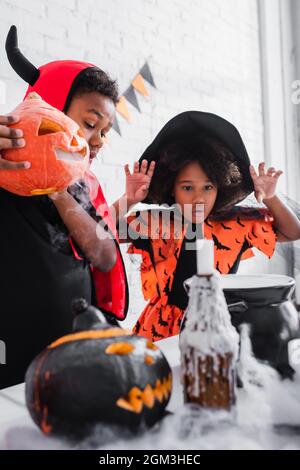 afroamerikanischer Junge im halloween Kostüm, der geschnitzten Kürbis hält, während er den Trank in der Nähe der gruseligen Schwester im spitzen Hut vorbereitet Stockfoto