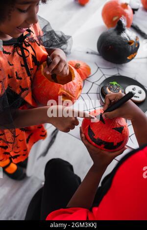 Glückliches afroamerikanisches Mädchen, das auf das Zeichnen auf Kürbis in der Nähe des Bruders zeigt Stockfoto
