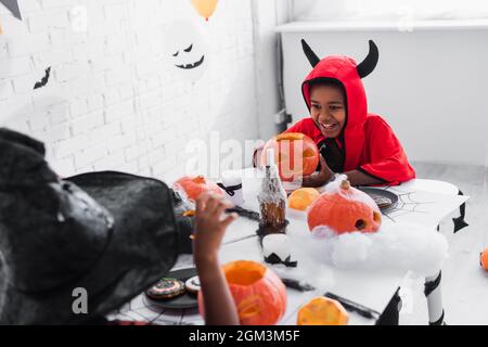 Glücklicher afroamerikanischer Junge im halloween Kostüm, der geschnitzten Kürbis in der Nähe verschwommener Schwester in spitzem Hut hält Stockfoto