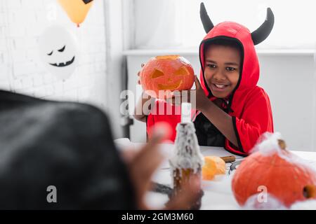 Glücklicher afroamerikanischer Junge in halloween Kostüm mit geschnitztem Kürbis in der Nähe verschwommener Schwester Stockfoto