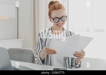 Glücklich lächelnde kaukasische Frau, die gute Nachrichten in Finanzdokumenten liest, während sie das Familienbudget verwaltet Stockfoto