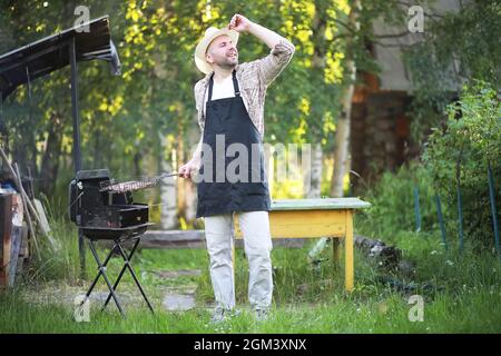 Grill Zeit. Europäischen Mann in hat Warten auf Gegrilltes. Er sitzt in der Nähe von Feuer und anderen. Stockfoto
