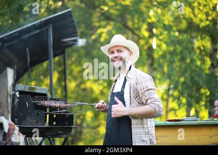 Grill Zeit. Europäischen Mann in hat Warten auf Gegrilltes. Er sitzt in der Nähe von Feuer und anderen. Stockfoto