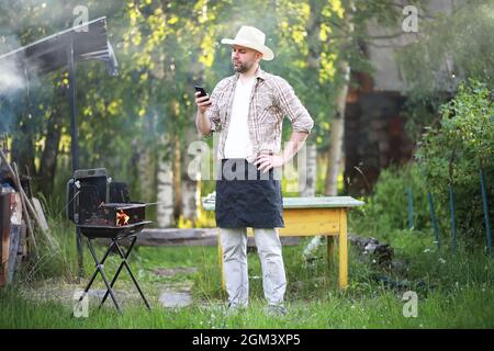 Grill Zeit. Europäischen Mann in hat Warten auf Gegrilltes. Er sitzt in der Nähe von Feuer und anderen. Stockfoto