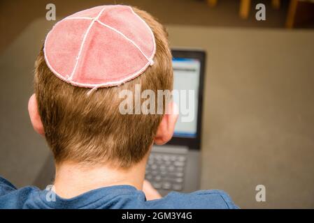 Kleiner Junge, der einen Yarmulke von hinten trägt und auf einem Laptop in einer Bibliothek mit bunten Büchern in den Regalen arbeitet. Stockfoto