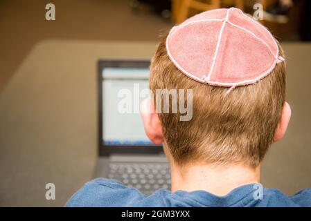 Kleiner Junge, der einen Yarmulke von hinten trägt und auf einem Laptop in einer Bibliothek mit bunten Büchern in den Regalen arbeitet. Stockfoto