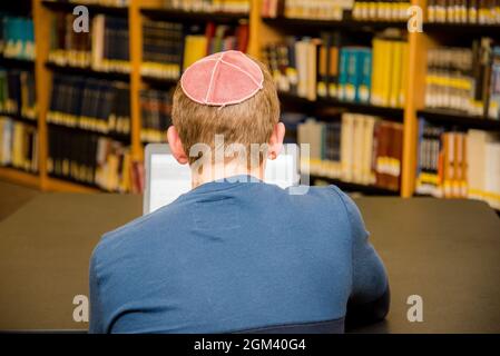 Kleiner Junge, der einen Yarmulke von hinten trägt und auf einem Laptop in einer Bibliothek mit bunten Büchern in den Regalen arbeitet. Stockfoto