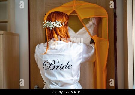 Rothaarige Frauen mit Blumenkranz in weißem Satingewand mit Aufschrift „Braut“ ziehen das Hochzeitskleid vom Kleiderbügel ab. Braut bereitet sich auf Hochzeit d Stockfoto