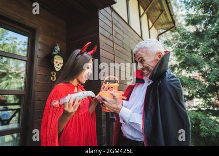 Aufgeregt reifer Mann in Vampir halloween Kostüm geben Eimer Süßigkeiten zu staunenden Tochter Stockfoto