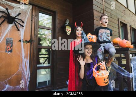 Mädchen im Hexenhut winkende Hand nahe aufgeregt Kinder in gruseligen halloween-Kostümen auf der Veranda mit Dekoration Stockfoto