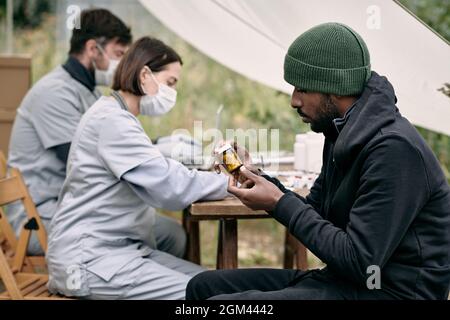 Serious junge schwarze Flüchtling Mann im Hut sitzt am Tisch der Mitarbeiter im Gesundheitswesen und lesen Tag der Pille Flasche, während sie kostenlose Medizin Stockfoto
