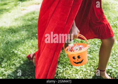 Verkürzte Ansicht eines Mädchens im roten Kostüm, das Halloween Eimer mit Süßigkeiten trägt, während es auf Gras läuft Stockfoto