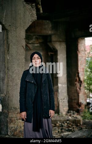 Porträt einer traurigen jungen muslimischen Flüchtlingsfrau in Hijab und langer Jacke, die gegen ein zerstörtes Gebäude steht Stockfoto