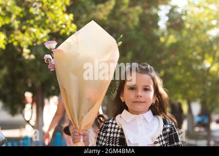 Ein kleines Mädchen mit langen dunklen Haaren in einer weißen Bluse und einer karierten Jacke hält einen Blumenstrauß in der Hand. Die erste Glocke der ersten Klasse in der Schule Stockfoto