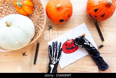 Auf einem von Kürbissen und Spinnen umgebenen Holzschreibtisch zeichnen Hände in schwarzen Handschuhen mit Knochen eine Skizze mit Markern eines lachenden Kürbisses, Draufsicht Stockfoto