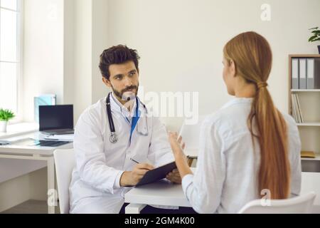 Aufmerksamer Arzt im Krankenhausbüro konsultiert weibliche Patienten und zeichnet Gesundheitsbeschwerden auf. Stockfoto