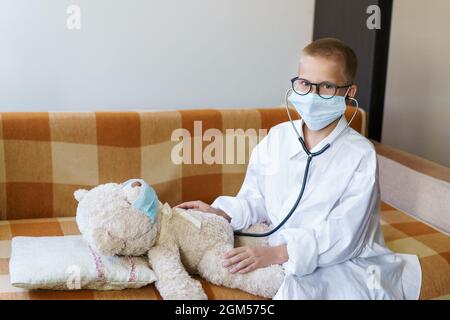 Niedliche kleine kaukasischen Jungen spielen lustige Spiel wie Arzt hält Stethoskop hören Vorschulkinder Spielzeug vorgibt, Krankenschwester zu sein behandeln flauschigen Patienten zu Hause. In einem Morgenmantel und einer medizinischen Maske. Stockfoto