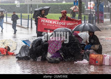Die Demonstranten vor der DSEI-Waffenmesse drängten sich während eines schweren Regensturms unter Regenschirme. Menschen, die sich vor Regen schützen. Banner der Friedensversprechen-Union Stockfoto