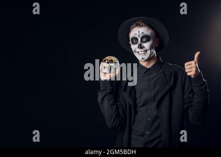 Junger Mann mit gemaltem Gesicht und Zuckerschädel für Mexikos Tag der Toten (El Dia de Muertos) auf dunklem Hintergrund Stockfoto