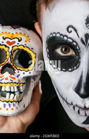 Junger Mann mit gemaltem Gesicht und Zuckerschädel für Mexikos Tag der Toten (El Dia de Muertos) auf dunklem Hintergrund, Nahaufnahme Stockfoto