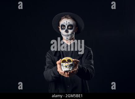 Junger Mann mit gemaltem Gesicht und Zuckerschädel für Mexikos Tag der Toten (El Dia de Muertos) auf dunklem Hintergrund Stockfoto