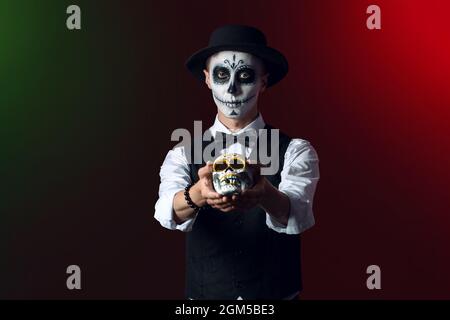 Junger Mann mit gemaltem Gesicht und Zuckerschädel für Mexikos Tag der Toten (El Dia de Muertos) auf dunklem Hintergrund Stockfoto
