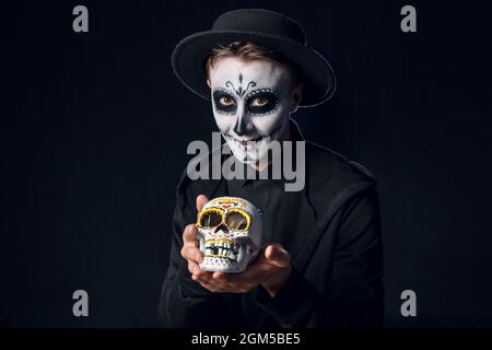 Junger Mann mit gemaltem Gesicht und Zuckerschädel für Mexikos Tag der Toten (El Dia de Muertos) auf dunklem Hintergrund Stockfoto