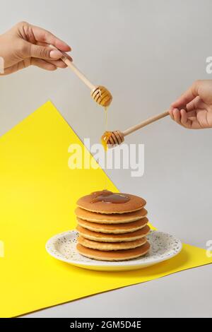 Weibliche Hände gießen Honig auf den Teller mit leckeren Pfannkuchen auf farbigem Hintergrund Stockfoto